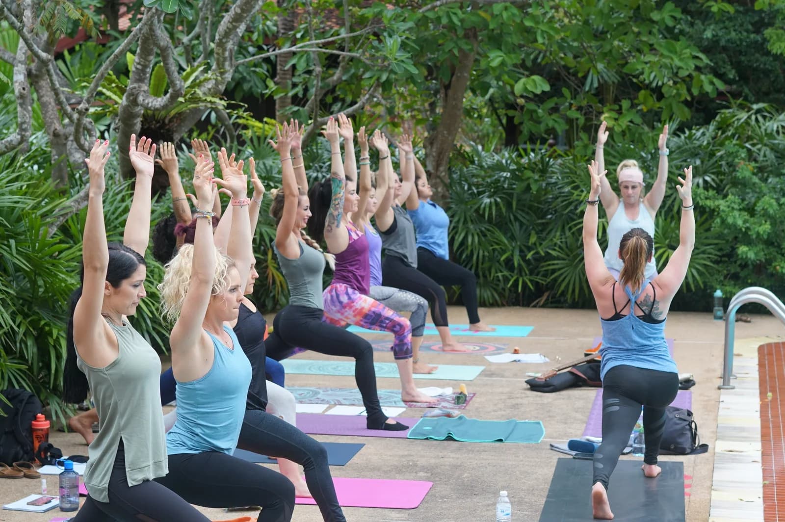 A session in the open air, everyone standing on their mats with both arms stretched overhead, jungle all around them.
